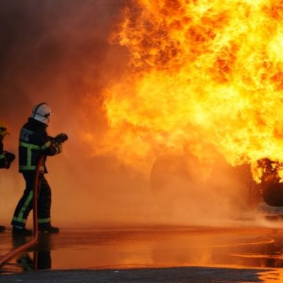 two people in helmets hold hoses and direct water spray at tall flames